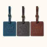 Three handcrafted square leather luggage tags in navy blue, dark coffee, and vintage brown with embossed travel quotes, brass buckle straps, and white stitching for travel bag identification.