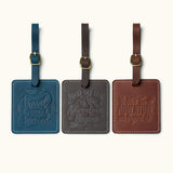 Three handcrafted square leather luggage tags in navy blue, dark coffee, and vintage brown with embossed travel quotes, brass buckle straps, and white stitching for travel bag identification.