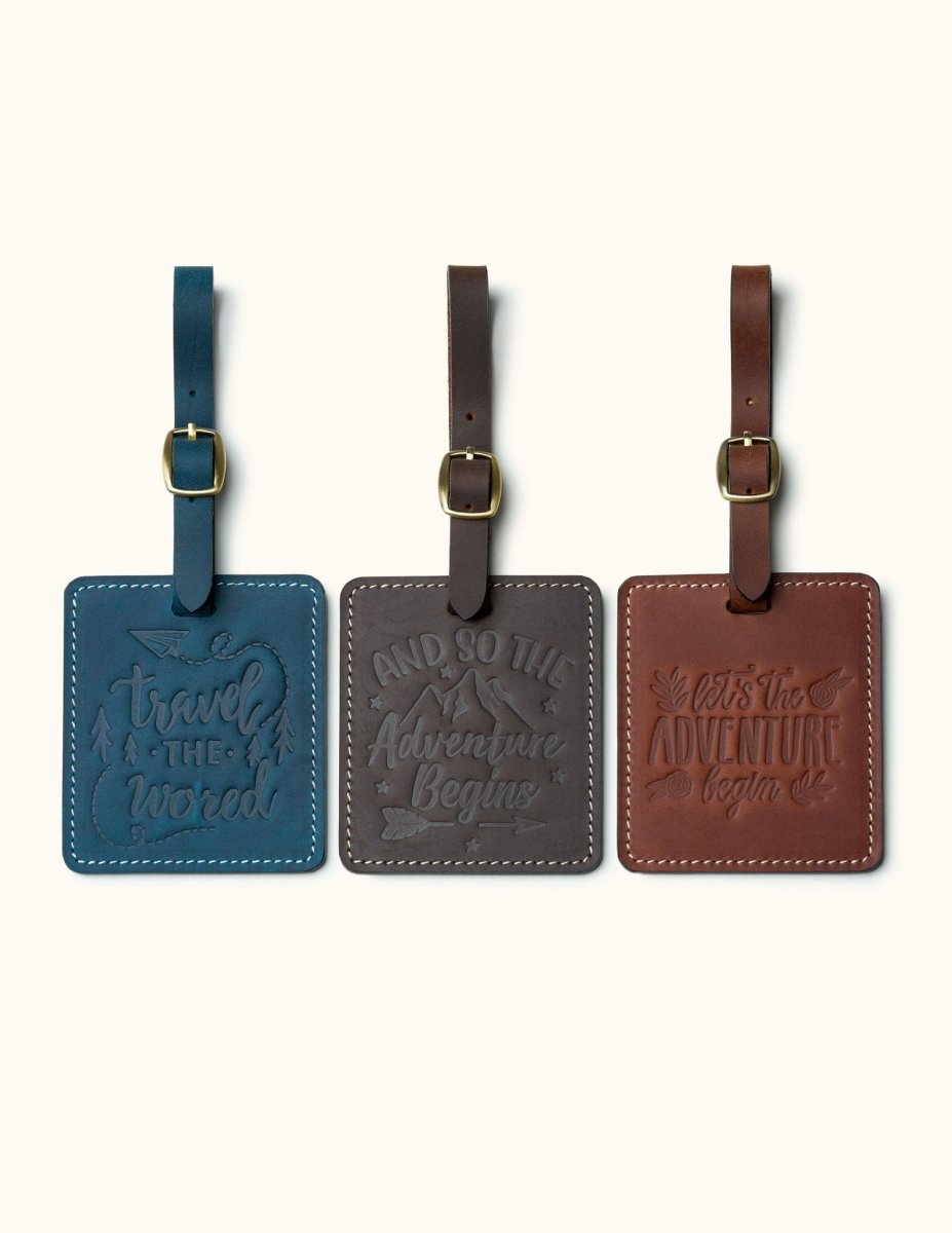 Three handcrafted square leather luggage tags in navy blue, dark coffee, and vintage brown with embossed travel quotes, brass buckle straps, and white stitching for travel bag identification.