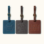 Three handcrafted square leather luggage tags in navy blue, dark coffee, and vintage brown with embossed travel quotes, brass buckle straps, and white stitching for travel bag identification.