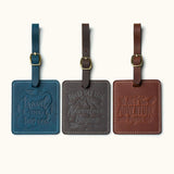 Three handcrafted square leather luggage tags in navy blue, dark coffee, and vintage brown with embossed travel quotes, brass buckle straps, and white stitching for travel bag identification.