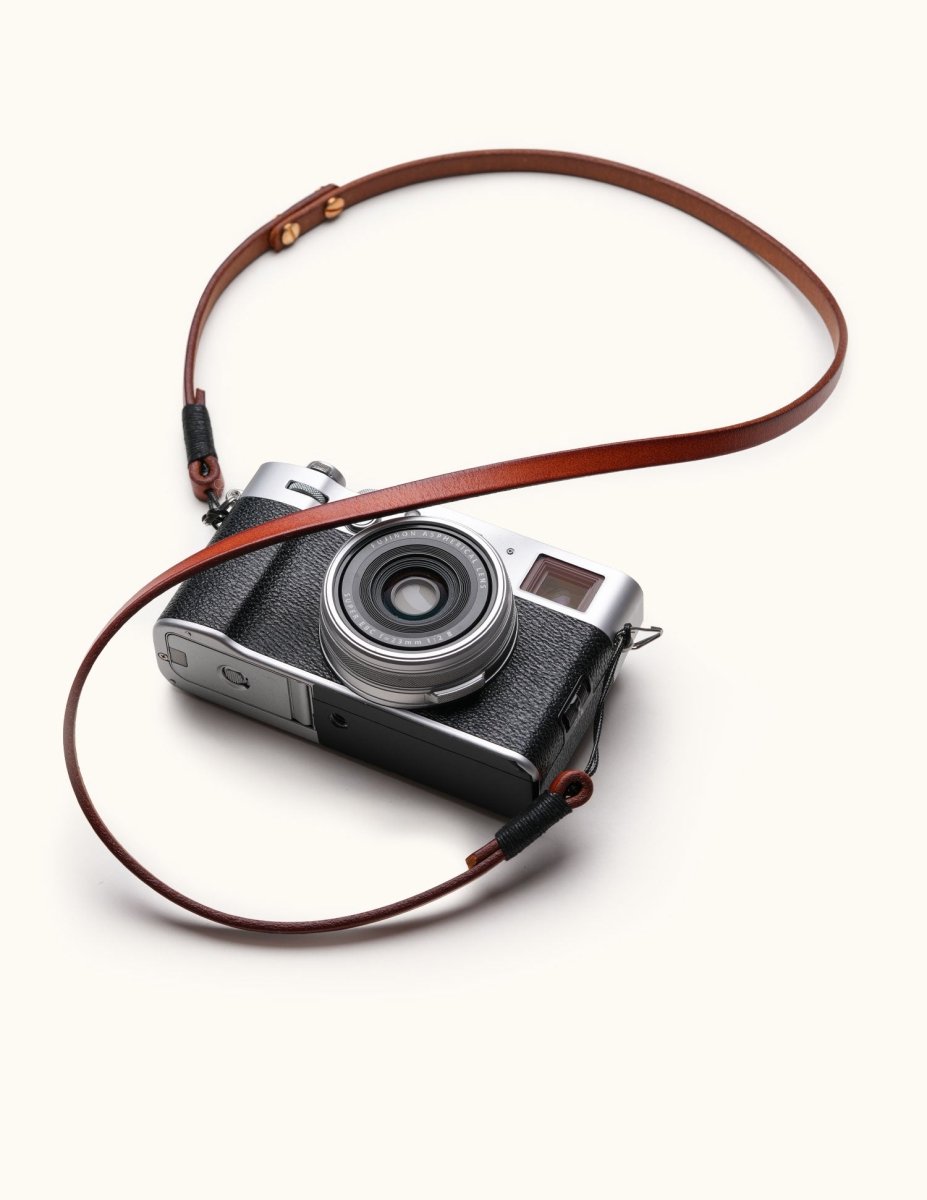 Classic full-grain leather camera strap attached to a Fujifilm mirrorless camera, shown in flat layout with visible wax cord and brass rivets