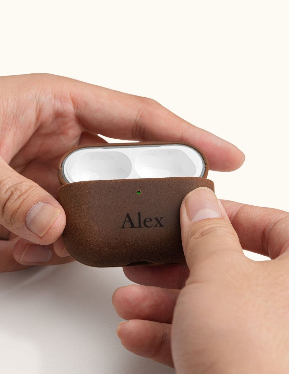 Hands holding an open vintage brown leather AirPods Pro case, displaying the earbuds inside. The front is custom laser engraved with the name Alex.