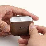 Hands holding an open vintage brown leather AirPods Pro case, displaying the earbuds inside. The front is custom laser engraved with the name Alex.
