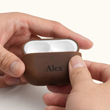 Hands holding an open vintage brown leather AirPods Pro case, displaying the earbuds inside. The front is custom laser engraved with the name Alex.