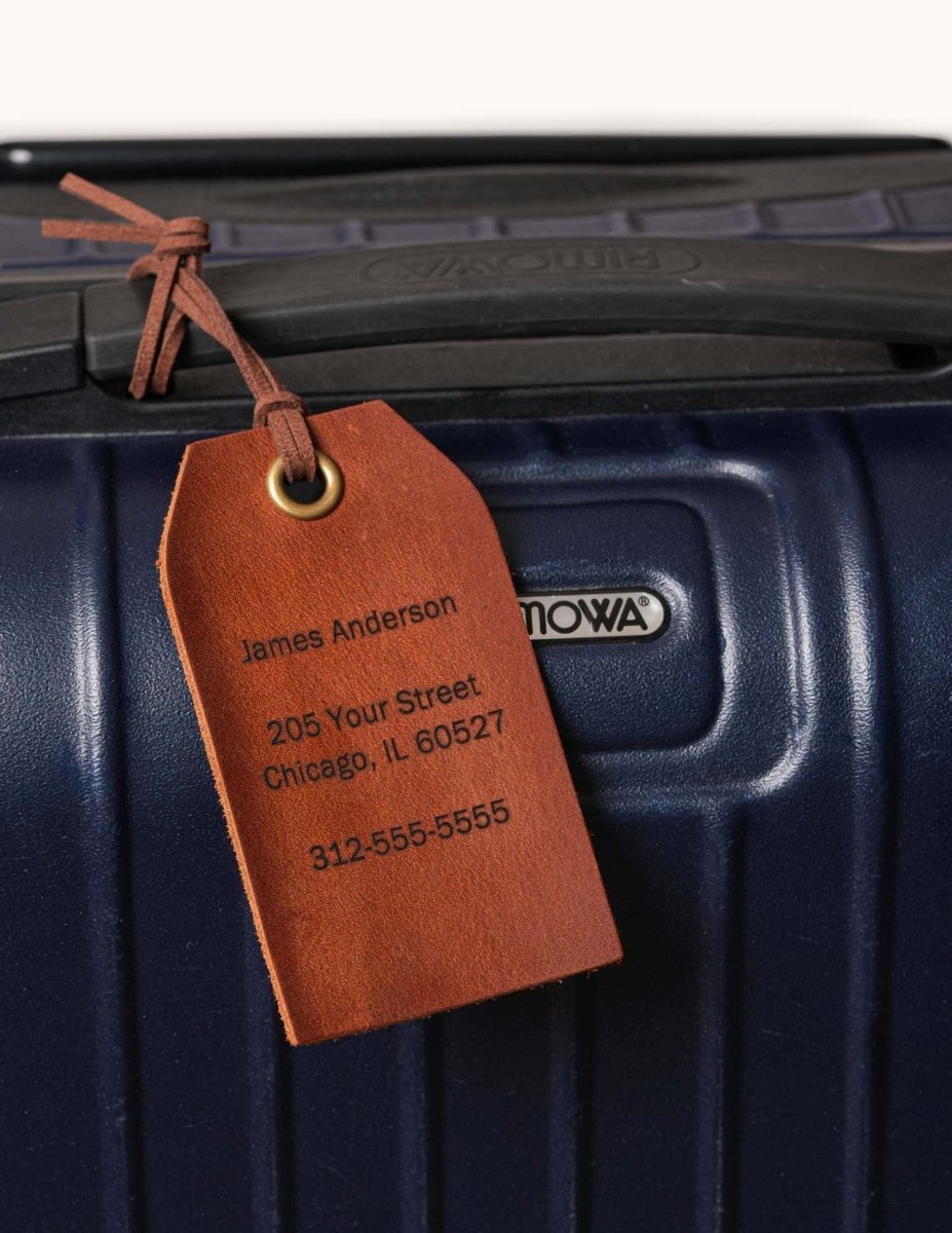 Personalized brown leather luggage tag attached to a navy suitcase handle, engraved with name, address, and phone number