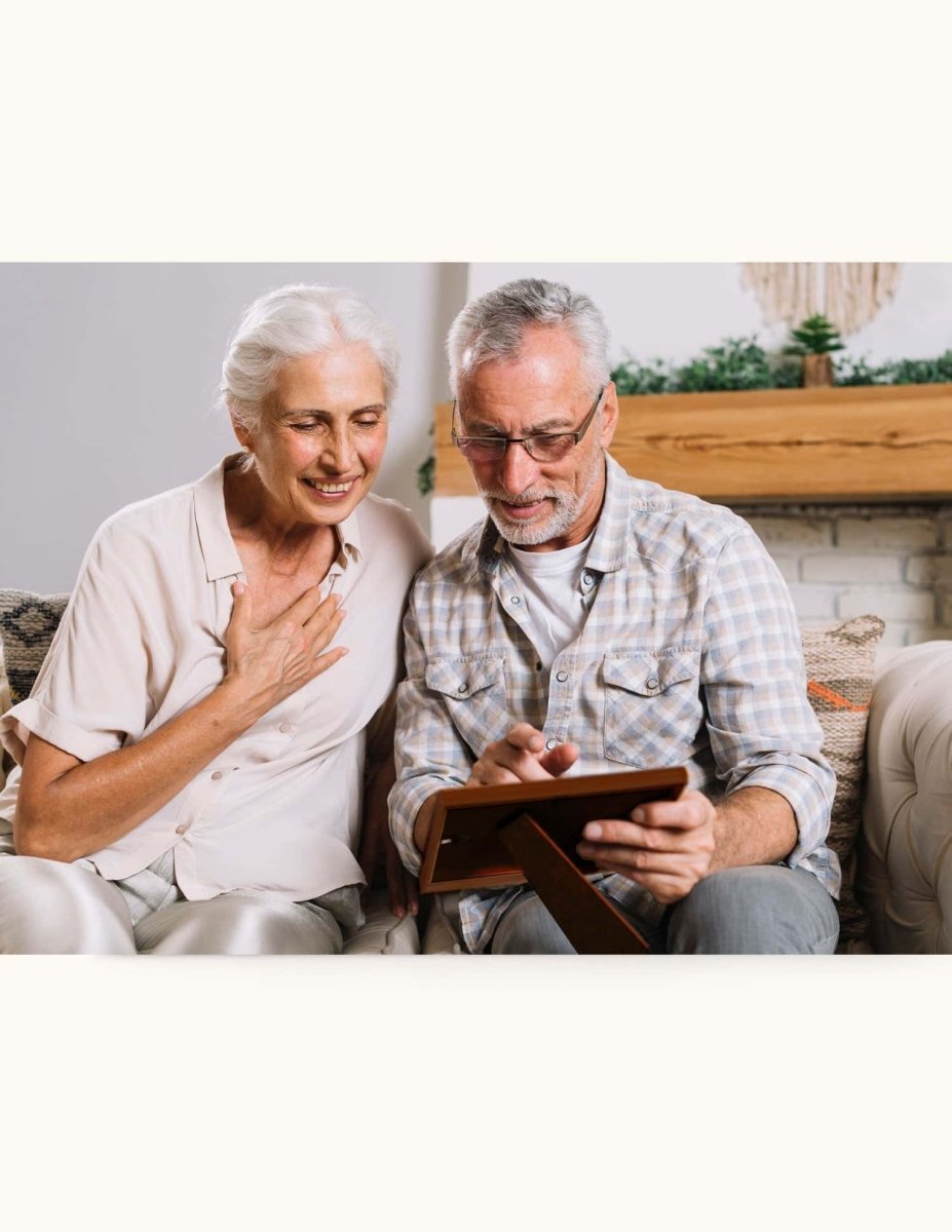Elderly couple sitting on a couch, emotionally touched while looking at a personalized photo frame — a meaningful anniversary gift filled with memories