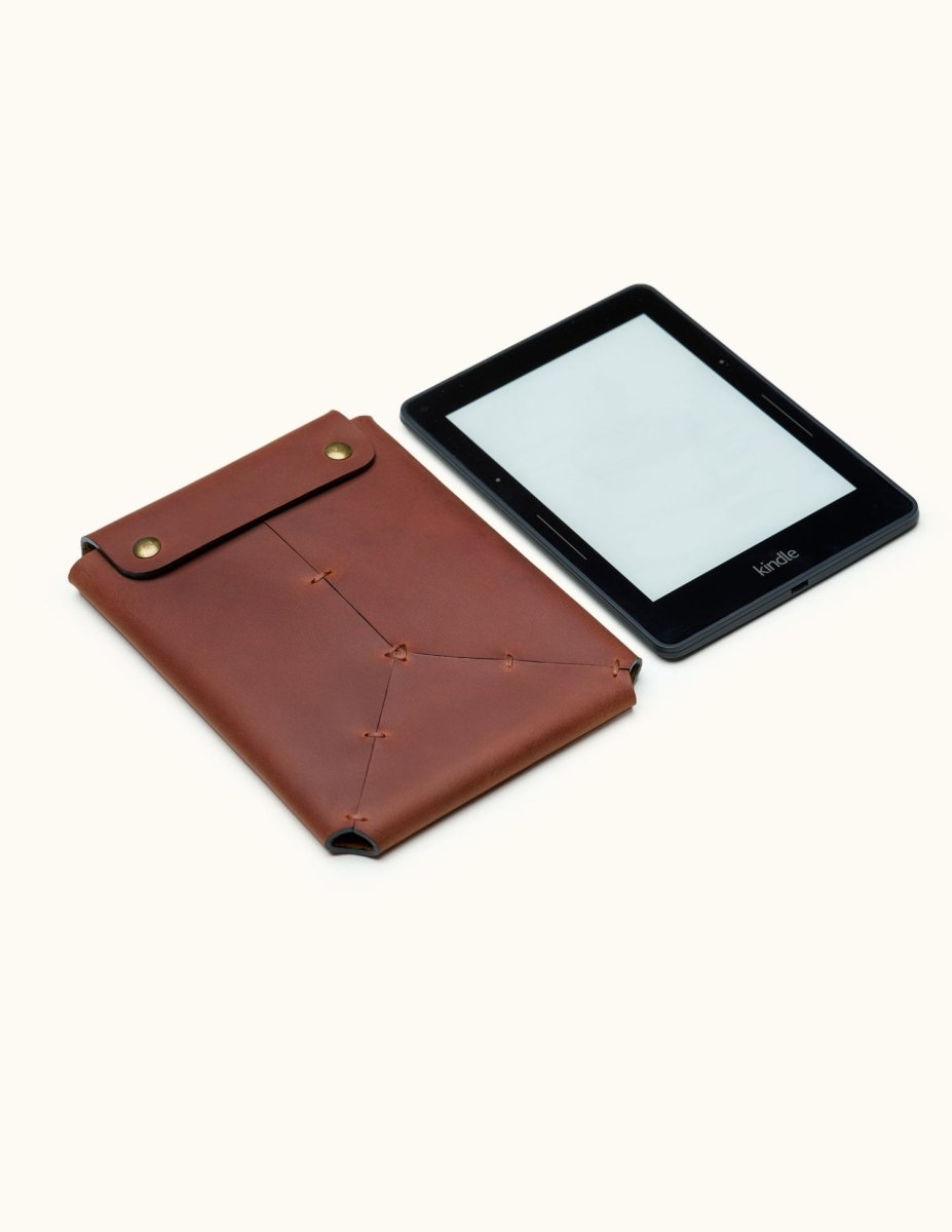 Genuine leather Kindle sleeve with brass snap buttons, shown next to a Kindle device. Hand-stitched design highlights premium craftsmanship