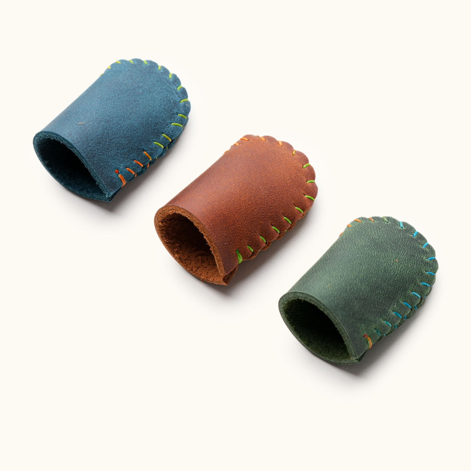 Three handmade full-grain leather thimbles in blue, brown, and green, used for finger protection during sewing or crafting.