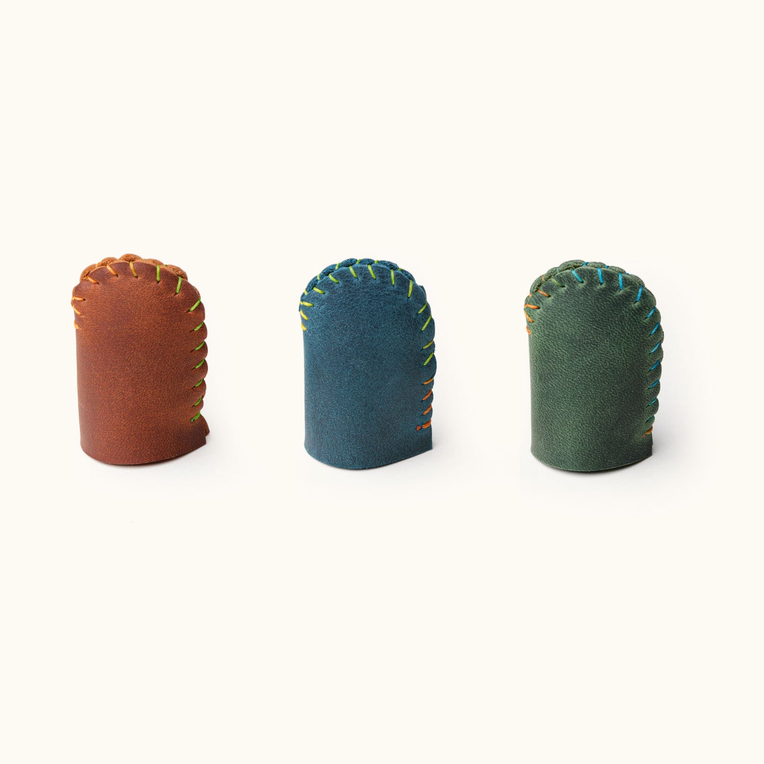 Three upright handmade leather thimbles in brown, blue, and green, shown standing to display their shape and stitching details.