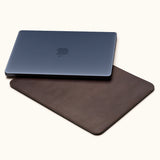 Closed dark blue laptop resting diagonally on a coffee brown leather sleeve against a cream background.