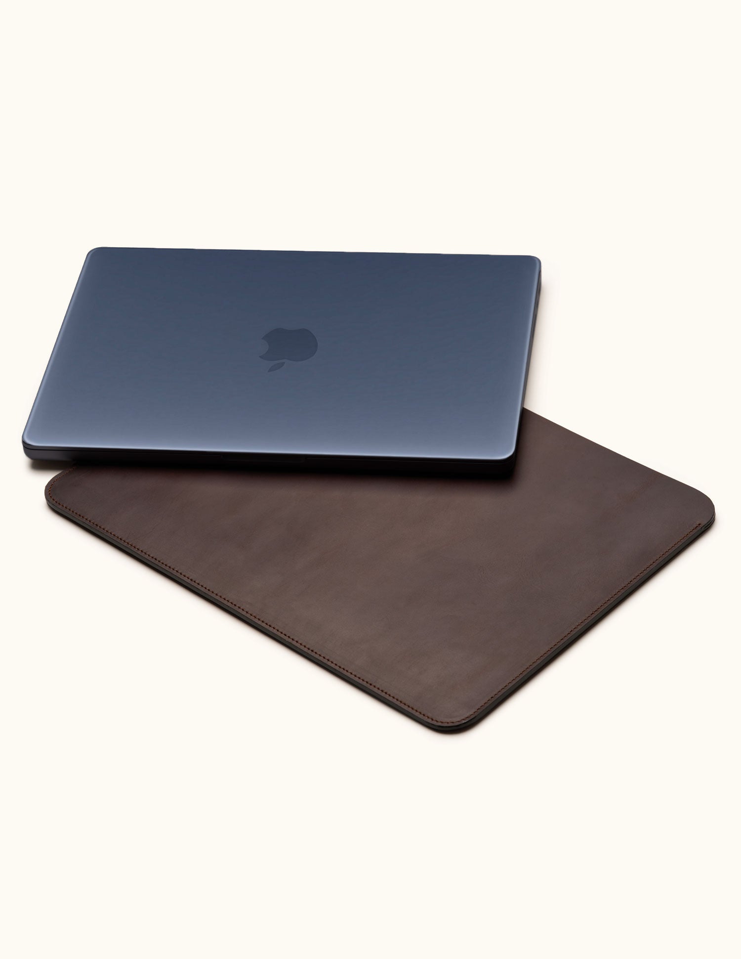 Closed dark blue laptop resting diagonally on a coffee brown leather sleeve against a cream background.