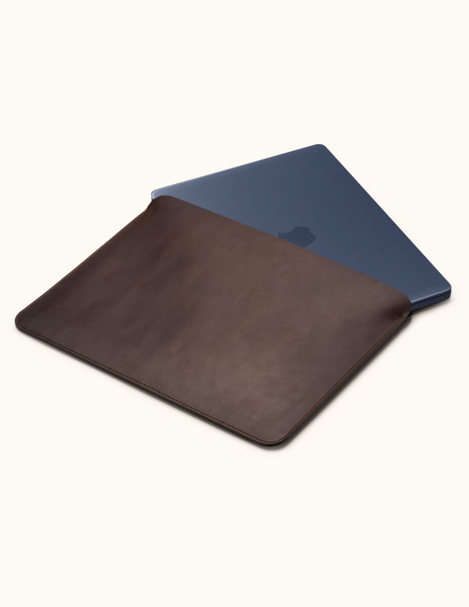 Coffee brown leather laptop sleeve with a dark blue laptop partially inserted, showing the open top and stitched edges.