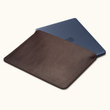 Coffee brown leather laptop sleeve with a dark blue laptop partially inserted, showing the open top and stitched edges.