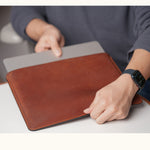 A person holding a vintage brown leather sleeve with a silver laptop partially inserted into the open top.