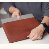 A person holding a vintage brown leather sleeve with a silver laptop partially inserted into the open top.