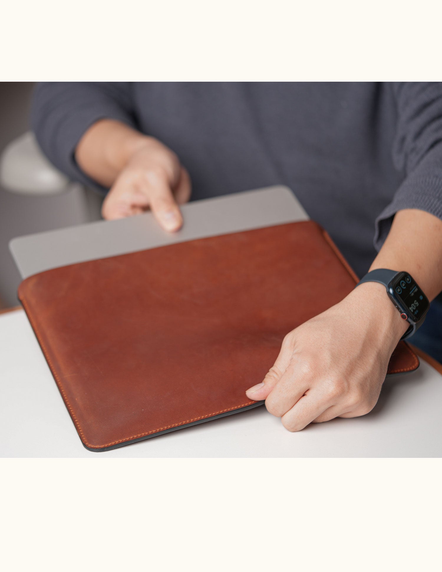 A person holding a vintage brown leather sleeve with a silver laptop partially inserted into the open top.