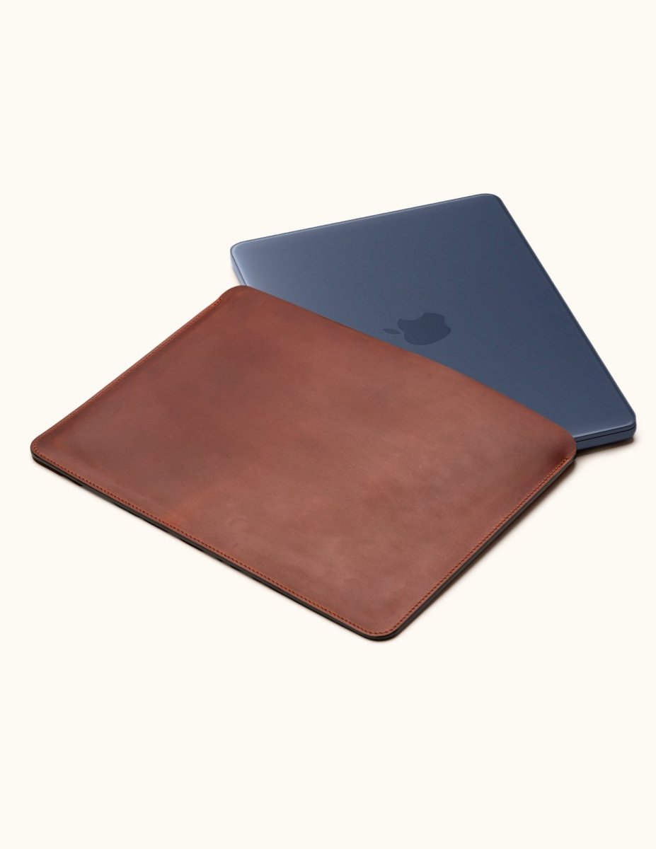 Vintage brown leather laptop sleeve with a dark blue laptop partially inserted, shown at an angle on a cream background.