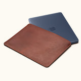 Vintage brown leather laptop sleeve with a dark blue laptop partially inserted, shown at an angle on a cream background.