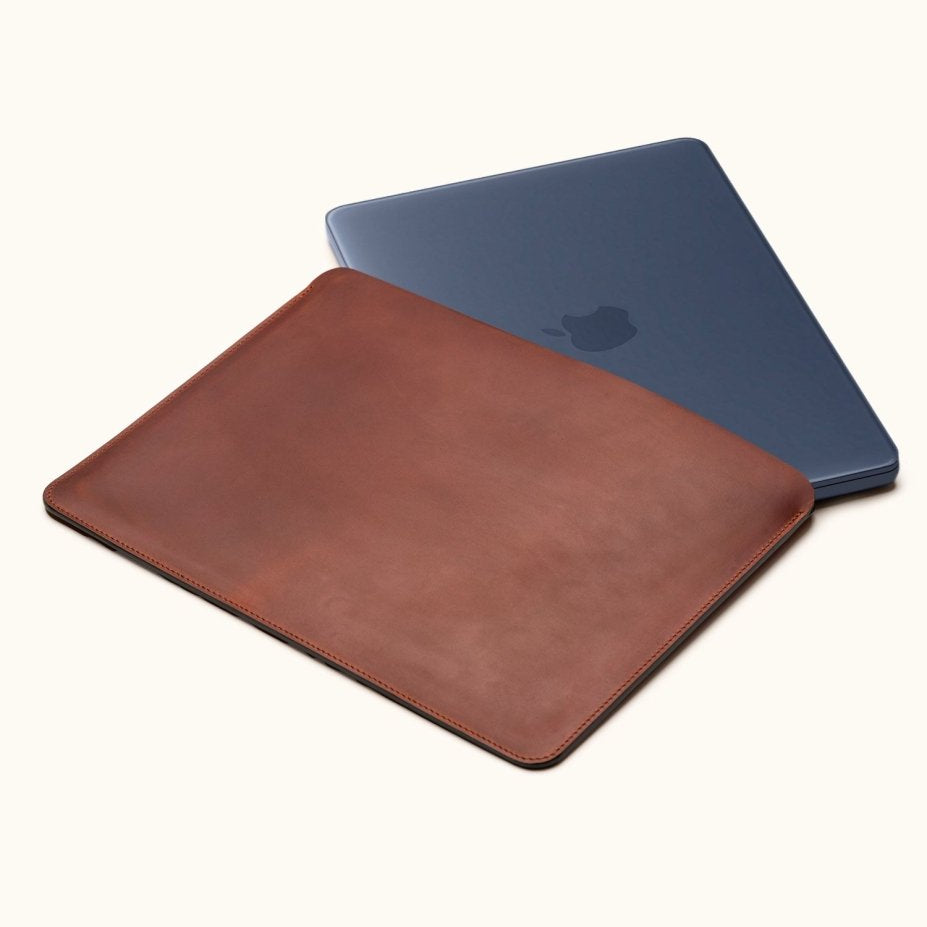 Vintage brown leather laptop sleeve with a dark blue laptop partially inserted, shown at an angle on a cream background.