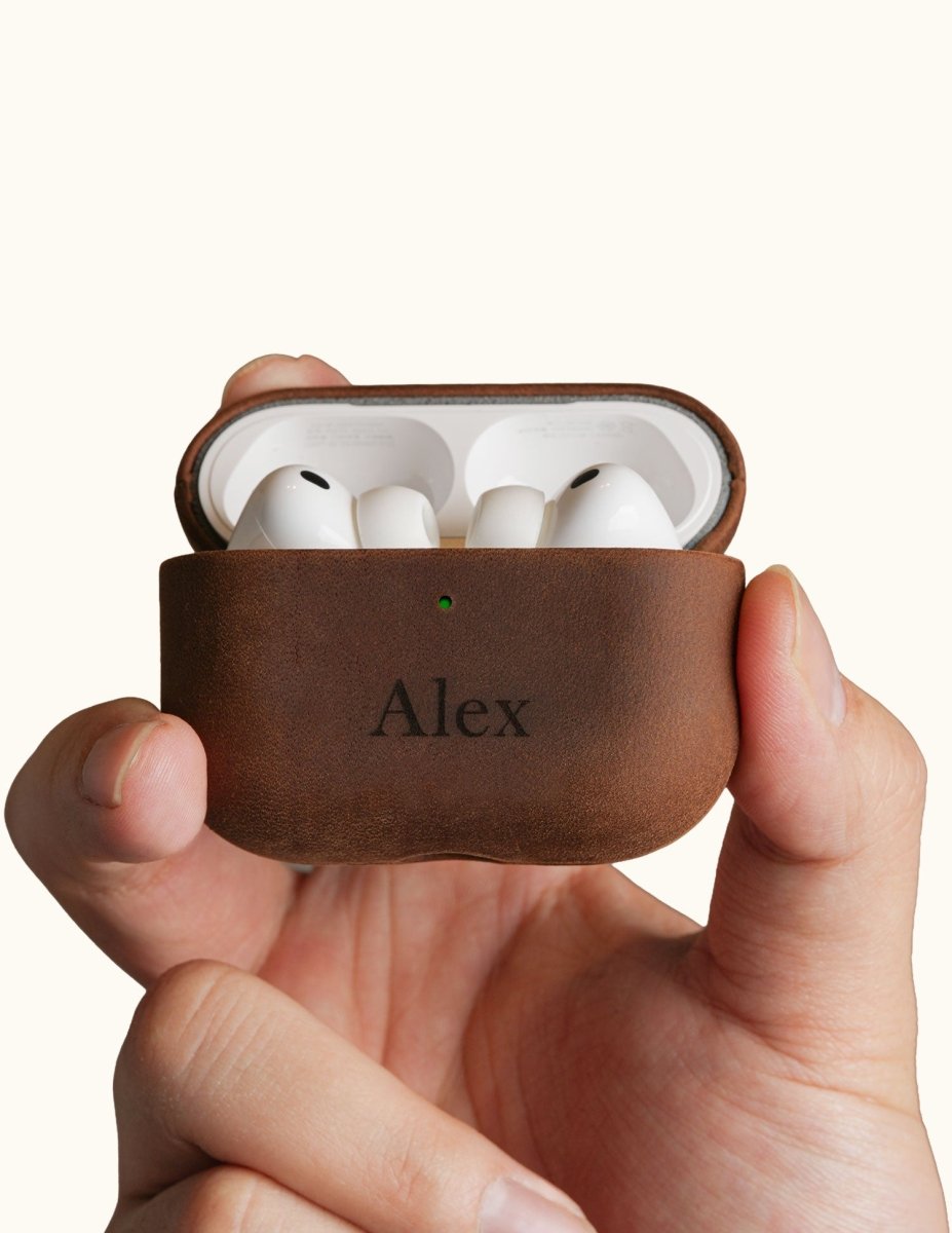 Hands holding an open vintage brown leather AirPods Pro case, displaying the earbuds inside. The front is custom laser engraved with the name Alex.
