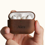 Hands holding an open vintage brown leather AirPods Pro case, displaying the earbuds inside. The front is custom laser engraved with the name Alex.