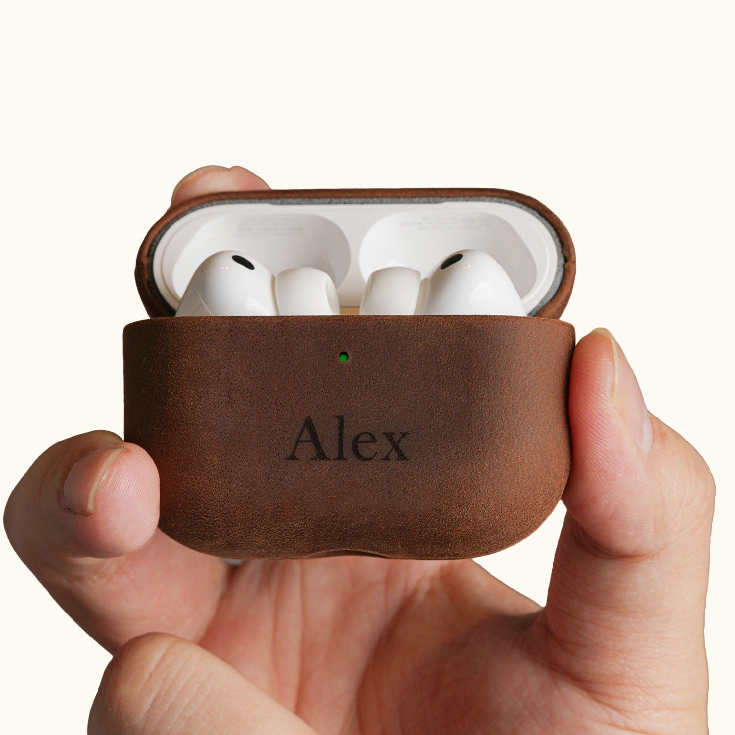 Hands holding an open vintage brown leather AirPods Pro case, displaying the earbuds inside. The front is custom laser engraved with the name Alex.