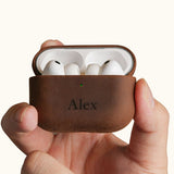 Hands holding an open vintage brown leather AirPods Pro case, displaying the earbuds inside. The front is custom laser engraved with the name Alex.