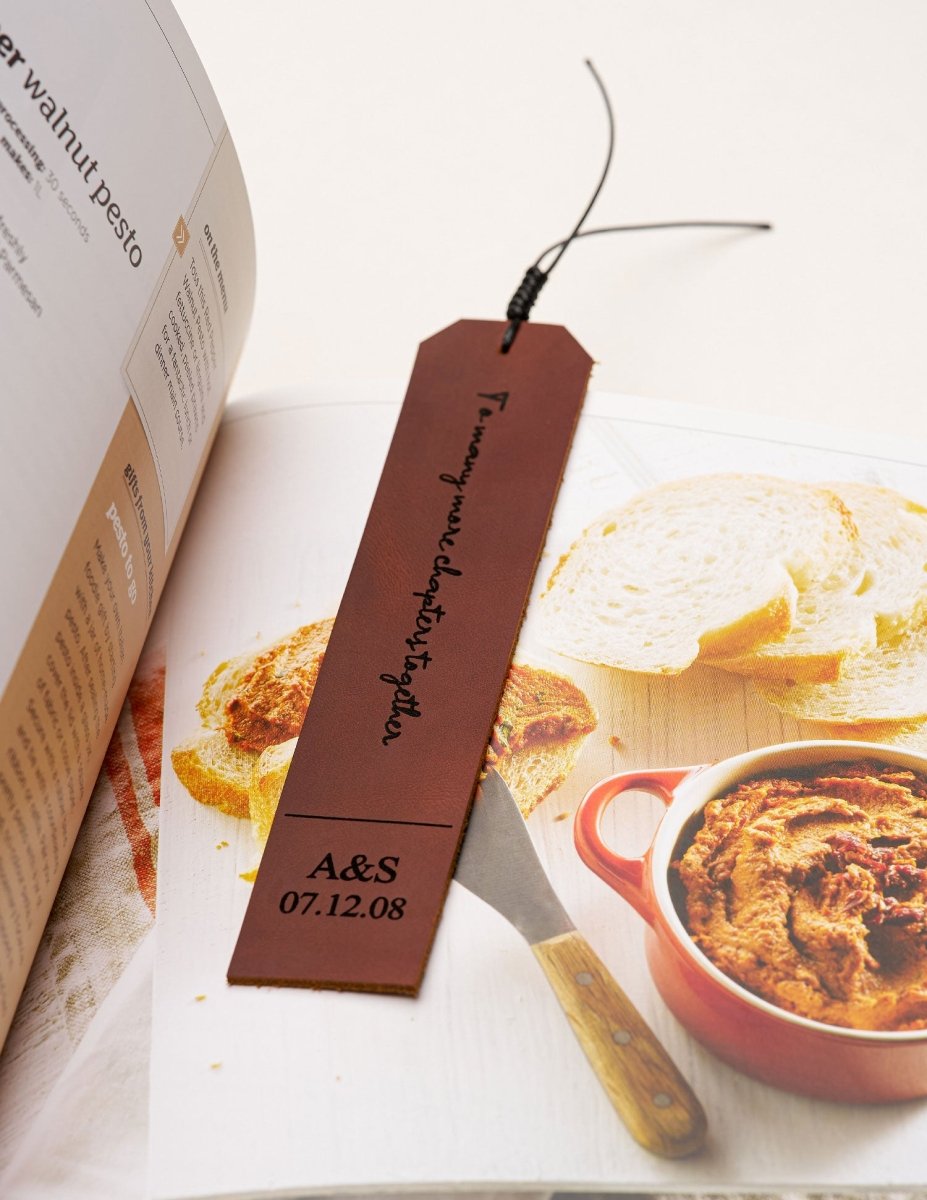Brown personalized leather bookmark engraved with "To many more chapters together" and a custom date, placed inside an open cookbook next to slices of bread and a red dish