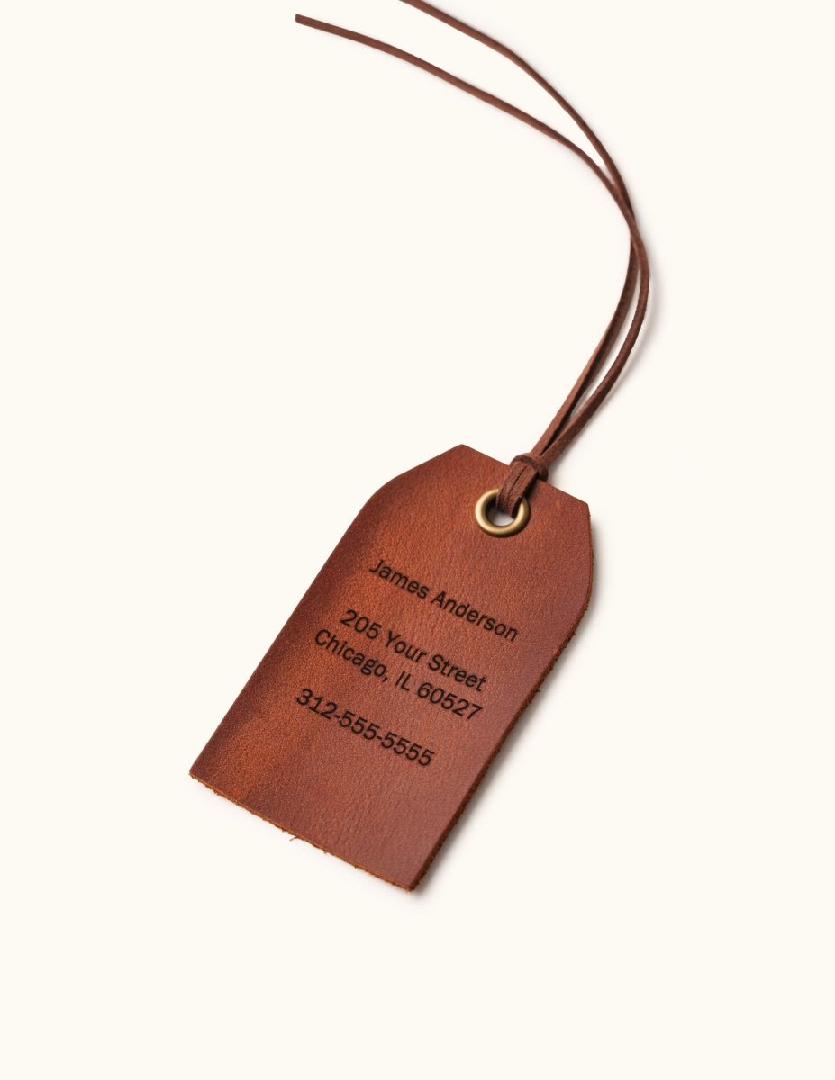 Personalized brown leather luggage tag engraved with the name James Anderson, address, and phone number, featuring a brass eyelet and leather strap