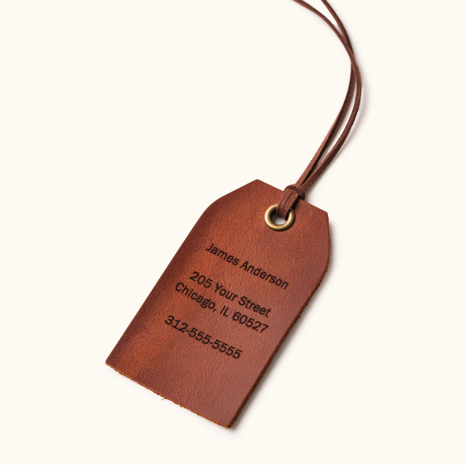 Personalized brown leather luggage tag engraved with the name James Anderson, address, and phone number, featuring a brass eyelet and leather strap