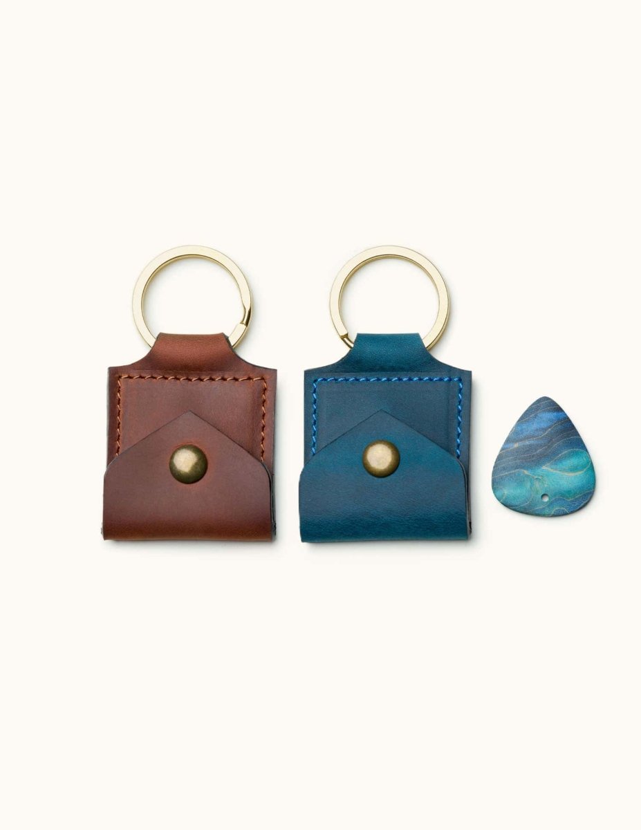Two square leather guitar pick holder keychains in brown and blue, shown with a pick for size reference